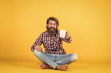 Vahşi sakallı adam kareli gömlek giyiyor, gür sakallı ve bıyıklı sabah kahvesini fincandan içiyor..