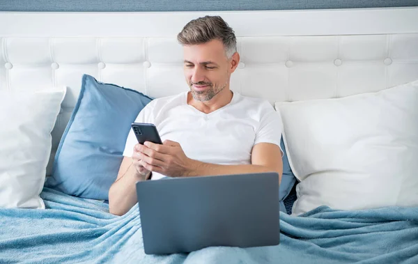Smiling Mature Man Chatting Phone Working Laptop Bed —  Fotos de Stock