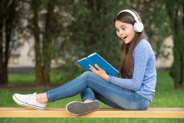 concept of teenager. Happy girl teenager reading and listening to audiobook in headphones after school outdoors, teenager.