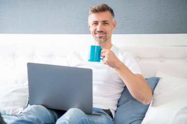 happy mature man using computer in bed with coffee.