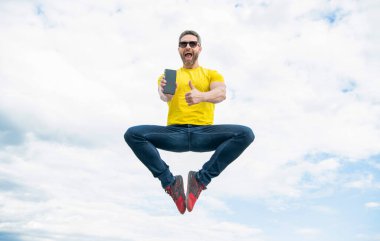 man jumping and presenting smartphone on sky background. thumb up.