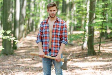Unshaven man in lumberjack style standing with axe forest background.
