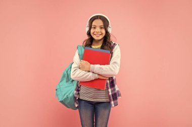 Okul çantasıyla kulaklıkla müzik dinleyen mutlu çocuk ve pembe arka plan, kitap çalışma kitabı..