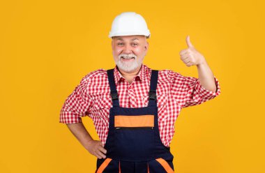 happy aged man builder in helmet on yellow background. thumb up.