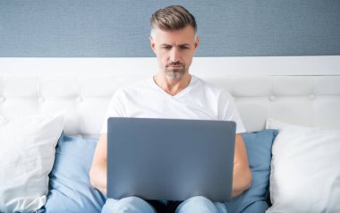 mature freelancer working on laptop in bed.