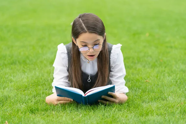 Surprised girl reading book lying on grass, reading. Teen girl reading outdoors.