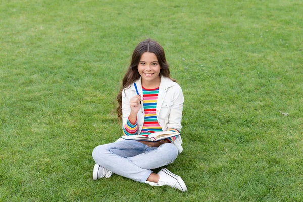 smiling teen girl making notes in notebook sitting on grass. taking notes. student make notes outdoor.