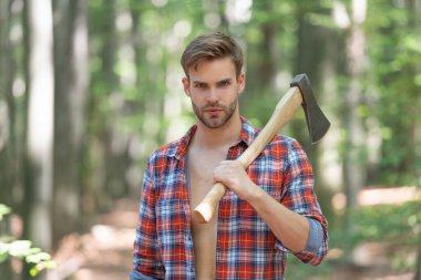Unshaven guy in lumberjack shirt holding axe on shoulder forest background.