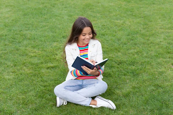 happy teen girl making notes in notebook sitting on grass. taking notes. student make notes outdoor.