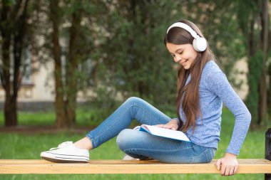 concept of audiobook. Happy teenage girl reading and listening to audiobook in headphones after school outdoors, audiobook.