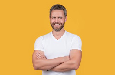 Happy man yellow studio background. Grizzled man smiling in casual white tshirt. Handsome man keeping arms crossed.
