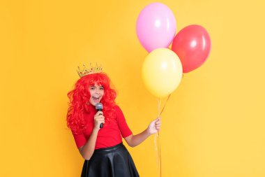 positive child in crown with microphone and party balloon on yellow background.