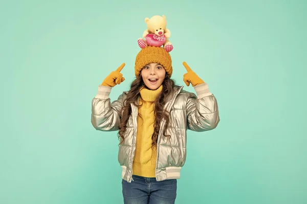 amazed child in hat and puffer jacket point finger on toy on blue background, toy shop.
