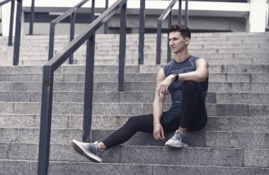 Kendine güvenen bir sporcunun portresi. Genç sporcu iyi bir koşudan sonra dışarıda oturuyor. Spor giyim bölümünde genç bir adam. Eğitimden sonra rahatla. Adam merdivenlerde oturuyor. Düşünceli, güçlü adam oturuyor..