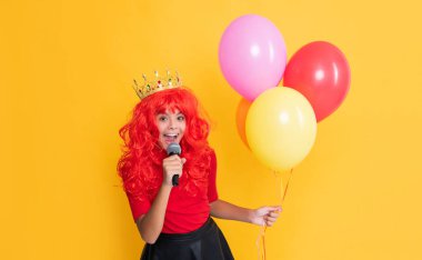 glad child in crown with microphone and party balloon on yellow background.
