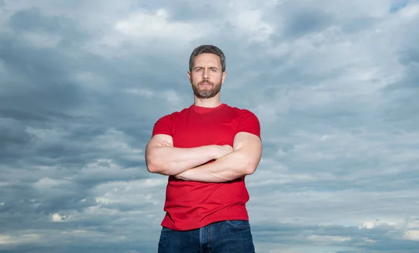 Serious man keeping arms crossed. Confident man sky background. Grizzled man in casual outdoors.