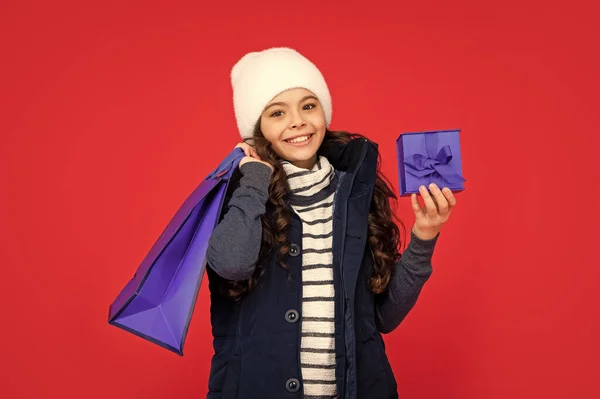 cheerful kid in puffer jacket and hat. teen girl after shopping on red background. child holding shopping bag and gift box. winter sales.
