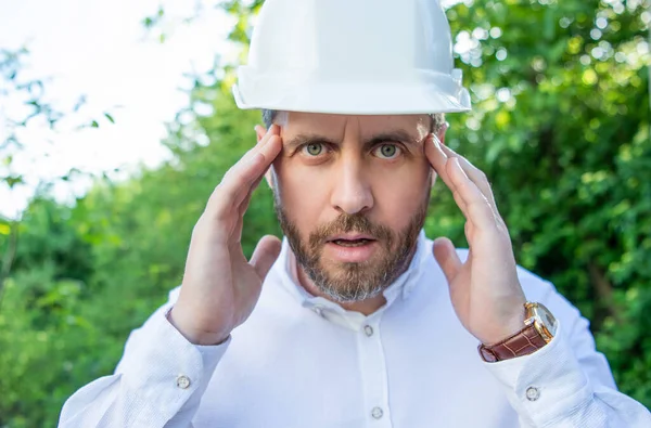 Shocked stressed businessman man in hardhat keeping hands at head outdoors.