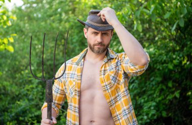 Serious cowboy man tipping cowboy hat holding pitchfork. Cowboy in garden natural background.