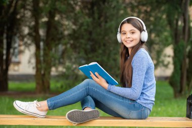 concept of teenage. Happy teenage girl reading and listening to audiobook in headphones after school outdoors, teenage.
