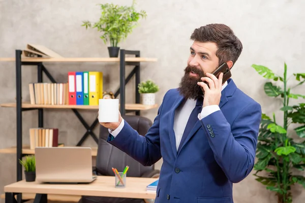 Happy professional man in suit talking on cellphone while drinking tea in office, phone.