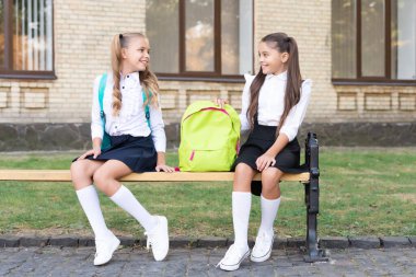 two school girls friends sit on bench with backpack together.