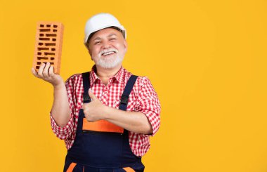 happy old aged man bricklayer in helmet on yellow background. thumb up.