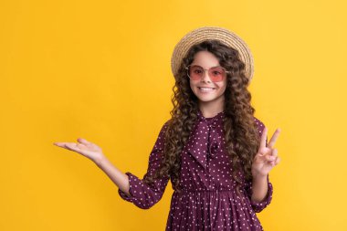 smiling child in straw hat and sunglasses with long brunette curly hair presenting product.