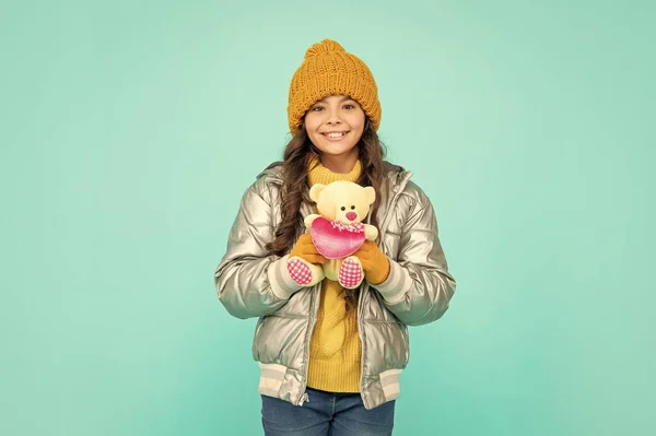 express positive emotion. winter fashion. love toy for valentines day. glad kid in puffer jacket and hat. childhood. teen girl hold toy bear. child wearing warm clothes on blue background.