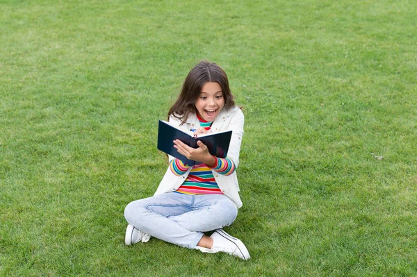 在笔记本上写笔记的少女坐在草地上惊讶极了 做笔记 学生在户外记笔记 — 图库照片