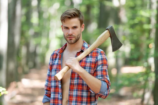 Lumbersexual man in lumberjack shirt holding axe on shoulder forest background.