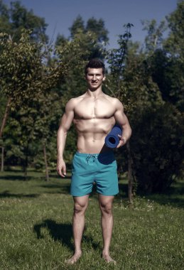 Atlet parkta elinde mavi yoga mat ile doğal peyzaj üzerinde güneşli yaz gününde gülümsemek. Spor, fitness, pilates. Sağlıklı yaşam konsepti