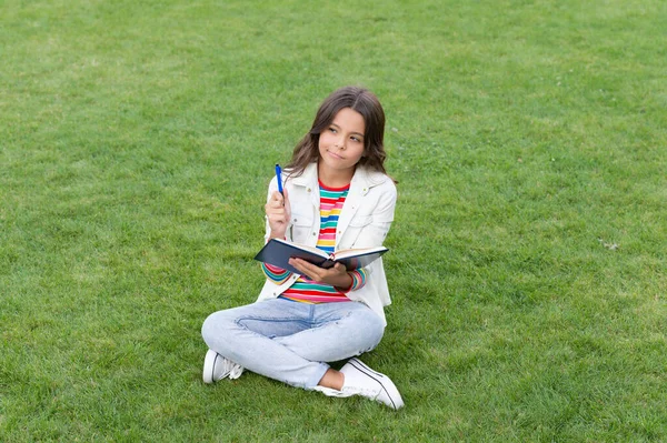 thoughtful teen child making notes in notebook sitting on grass. taking notes. student make notes outdoor.