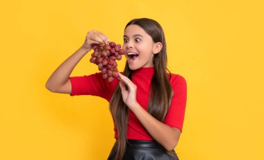 Şaşırmış çocuk sarı arka planda bir demet üzüm tutuyor.