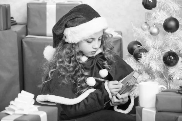 愿望清单 孩子们的桑塔服装享受圣诞节前夕 孩子们写信给圣塔克洛斯 亲爱的桑塔女孩在圣诞树旁和礼品盒旁拿着笔和纸 相信奇迹 发信给桑塔 — 图库照片