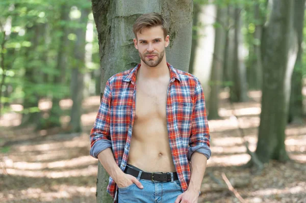 ハンサムな若いです男とともにLumbersexルックスでUnbuttoned Lumberjackシャツ立っている木の森の背景 — ストック写真