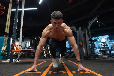 Çömelme pozisyonu almış, dizlerinin üstüne çökmüş ve elleri spor salonunda başlangıç çizgisinde. Koşmak için formda bir adam başlangıç pozisyonunda. Spor salonunda atlet eğitimi. Spor salonunda koşu antrenmanı. Spor ve zindelik.
