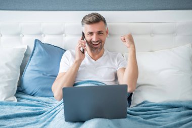 Başarılı bir yetişkin telefonda konuşuyor ve laptopla yatakta çalışıyor..