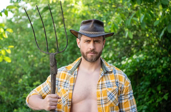Portrait of man in farmer hat. Serious farmer holding hayfork. Farmer in garden natural background.
