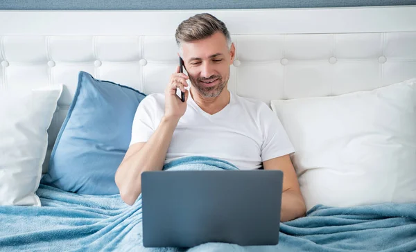 cheerful mature man speaking on phone and working on laptop in bed.