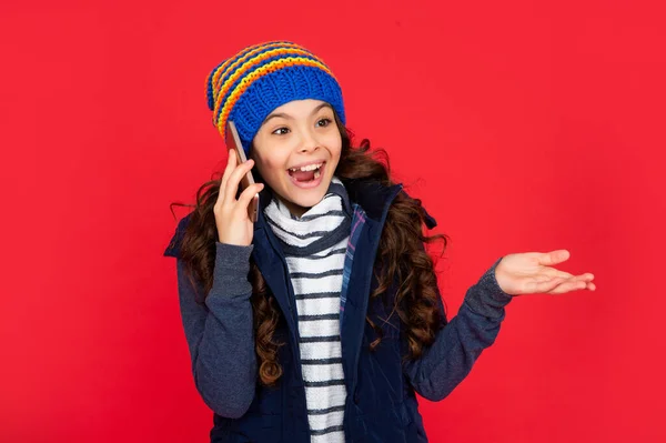 通信だ 幸せな冬の休日 赤い背景で帽子をかぶった子供が電話で話しているのに驚きました スマホでベストを着た10代の女の子 子供の挨拶や電話での会話の肖像 — ストック写真