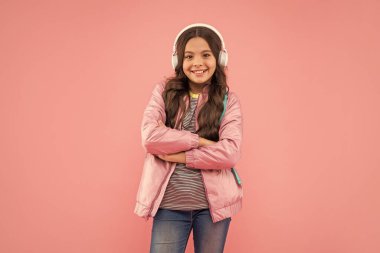cheerful kid listening music in earphones with school bag on pink background, music.