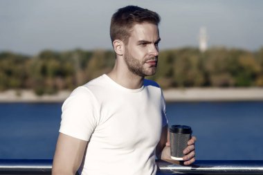 Basit bir yaz eğlencesi. Genç adam nehir kıyısında kahve içmekten hoşlanıyor. Yakışıklı adam sıcak bardağı dışarıda tutuyor. Sabah ayini. Kafein enerjisi. İç ve keyfini çıkar. Kahveden sonra hayatın tadını çıkar. Afiyet olsun..