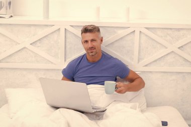 Awake man drink coffee using laptop computer in bed, morning.