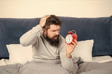 Perplexed guy scratching head holding alarm clock being in bed in morning, oversleep.