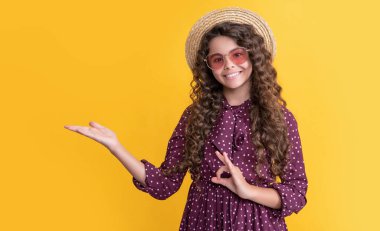 Hasır şapkalı, güneş gözlüklü, uzun kıvırcık saçlı neşeli çocuk ürününü sunuyor..