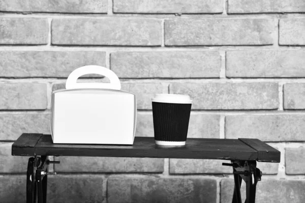 Take away coffee and white package on table. Food delivery and hot beverage concept. Cup of take away drink and food box on low table near stylish wall. Paper cup and container on brick background.