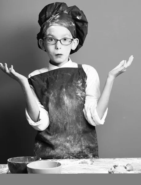 stained cute cook chef boy. young boy cute cook chef in uniform and hat on stained surprised face flour with glasses standing near table with molds for cookies, colorful bawls with spread hands on red