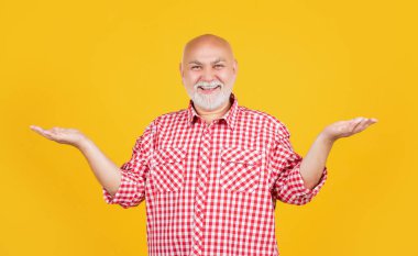 Kareli tişörtlü mutlu yaşlı adam sarı arka planda ürün sunuyor..