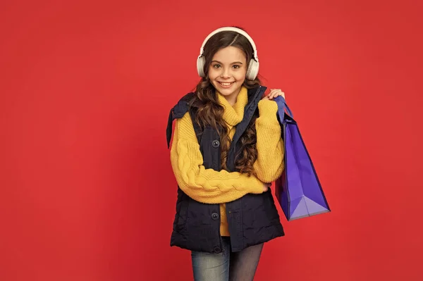 冬の帽子とイヤフォンの幸せな子供は赤い背景にショッピングバッグを保持し — ストック写真